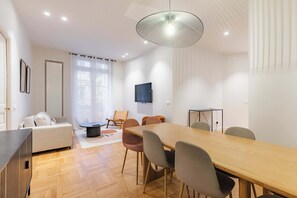 Ein modernes Wohnzimmer mit einer Couch, einem Esstisch mit Stühlen, einem an der Wand befestigten Fernseher und einem großen Fenster mit Vorhängen.