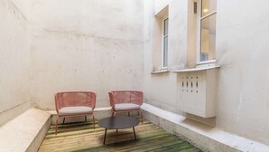 Ein kleiner, abgeschlossener Außenbereich mit zwei Korbstühlen und einem runden Tisch, einem Betonboden und einer weißen Wand mit einem Fenster.