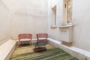 Ein kleiner, abgeschlossener Außenbereich mit zwei Korbstühlen und einem runden Tisch, einem Betonboden und einer weißen Wand mit einem Fenster.