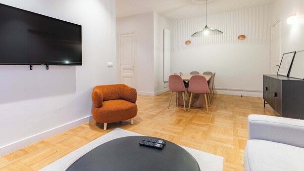 Ein modernes Wohnzimmer mit Flachbildfernseher, rundem Couchtisch, Sofa, Essbereich mit Tisch und Stühlen sowie einem Schrank.