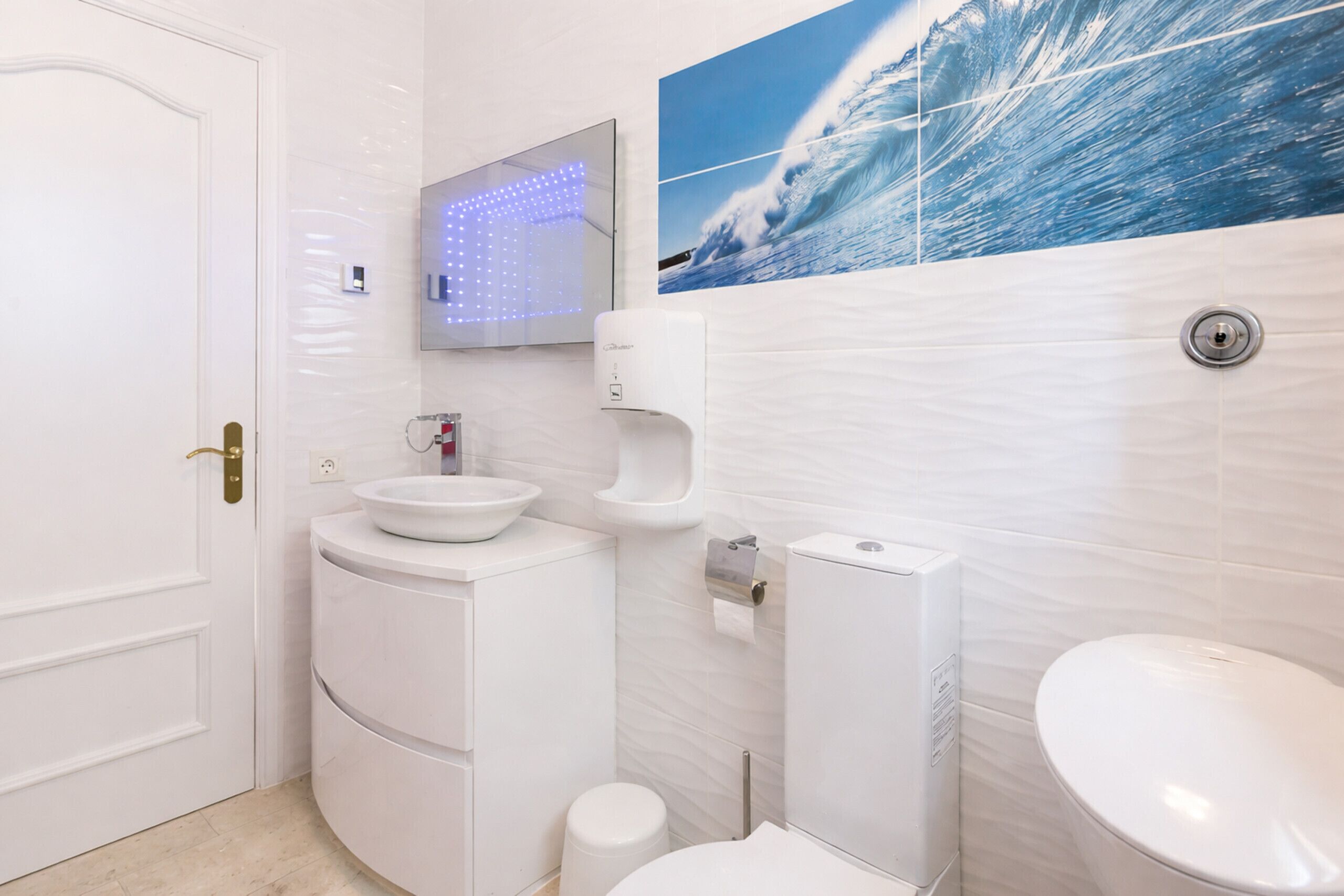 A modern bathroom with a white sink, toilet, and bidet, a mirror with LED lights, and a wave-themed wall art.