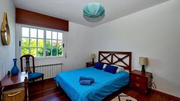 Une chambre avec une grande fenêtre, un lit avec une literie bleue, une tête de lit en bois, une table de chevet avec une lampe et une chaise.