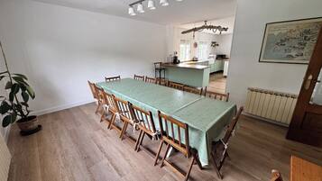 A dining area with a long table, chairs, a potted plant, and a kitchen in the background.
