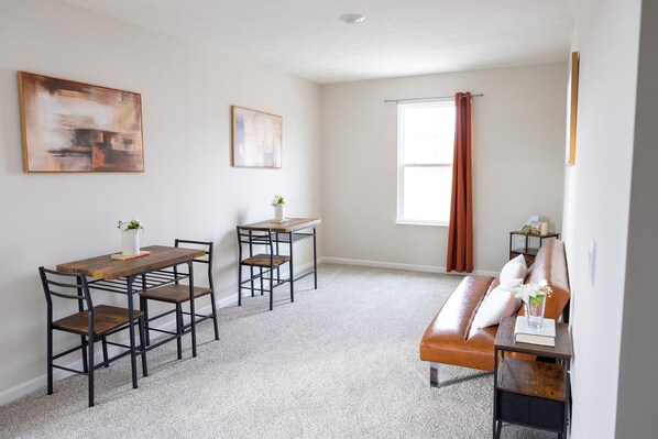 A modern living room with a brown leather sofa, a wooden dining table with metal chairs, and a small side table with a vase. - Holistic Campus Rental, LLC - Private Bedrooms in Shared Home with Shared Kitchen (Oxford)