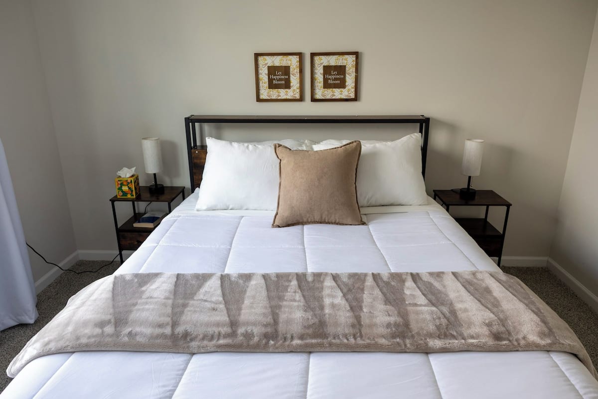 A neatly made bed with a beige throw blanket, two framed motivational quotes on the wall, and bedside lamps on either side.