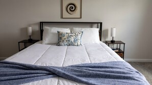A neatly made bed with a blue blanket, two bedside lamps, and a framed wall decoration. - Holistic Campus Rental, LLC - Private Bedrooms in Shared Home with Shared Kitchen (Oxford)