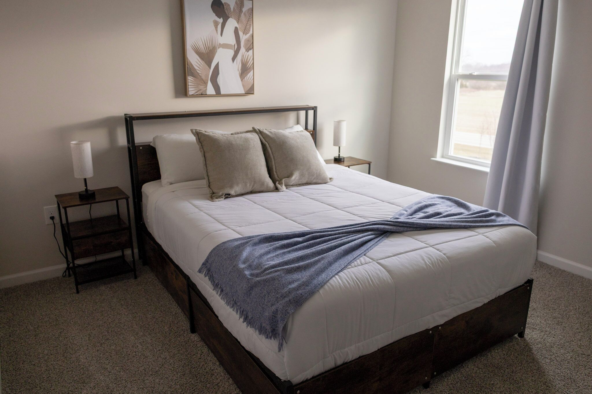 A neatly made bed with a white comforter and two pillows, a bedside table with a lamp, and a framed artwork above the bed.