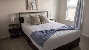 A neatly made bed with a white comforter and two pillows, a bedside table with a lamp, and a framed artwork above the bed.