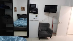 Un dormitorio con cama, un refrigerador blanco pequeño, un microondas, un sillón gris, un televisor y un armario.
