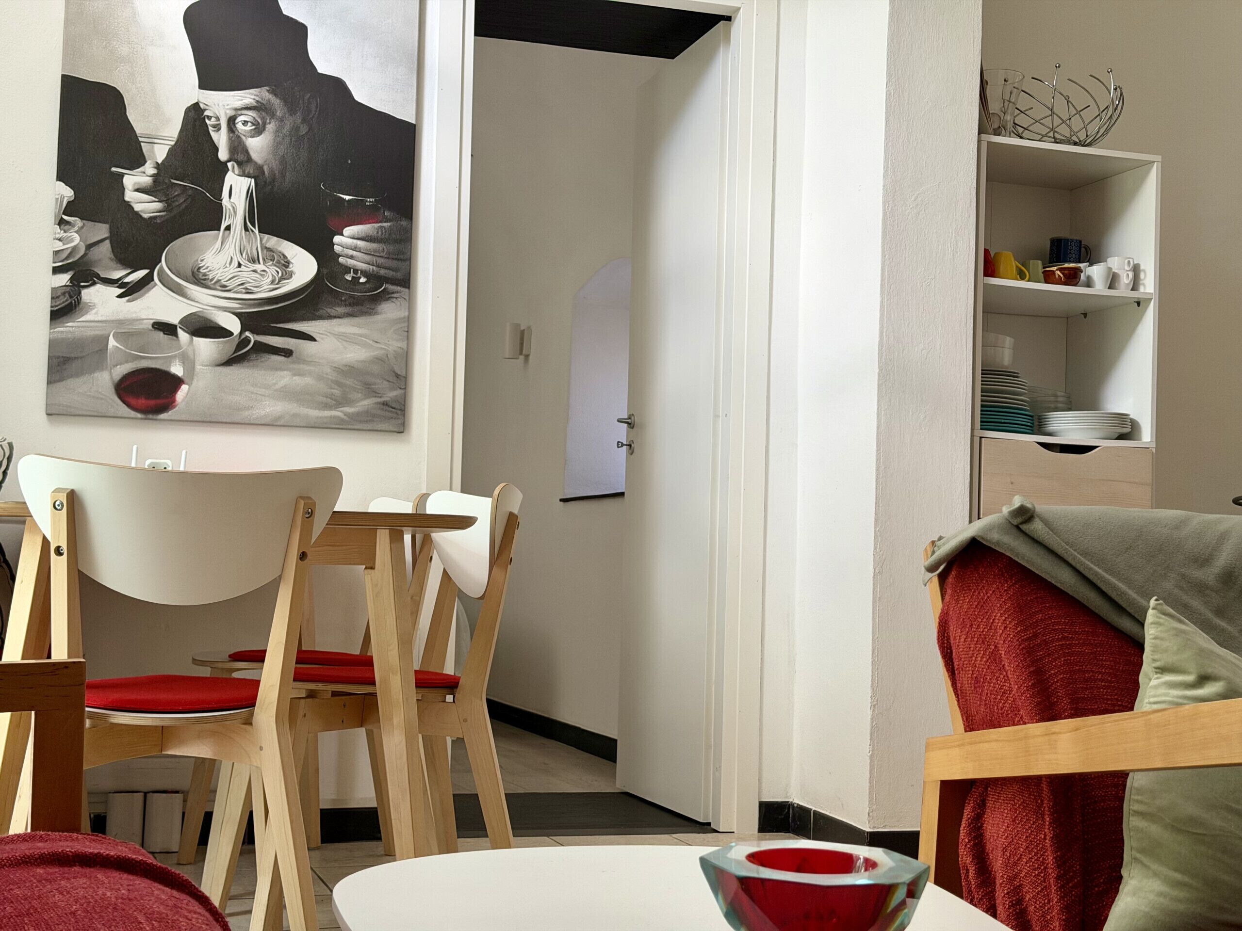 A dining area with a table, chairs, a painting, and a shelf with dishes.