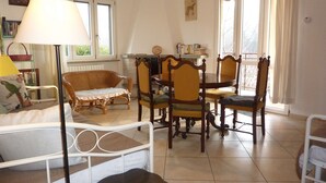 Een gezellige kamer met een bed, een eettafel met stoelen, een rieten fauteuil en een lamp.