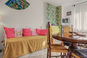 Ein Zimmer mit einem Bett, einem Tisch und einem Stuhl. Darüber hängt ein farbenfrohes Wandbild und eine Bücherwand mit Büchern.
