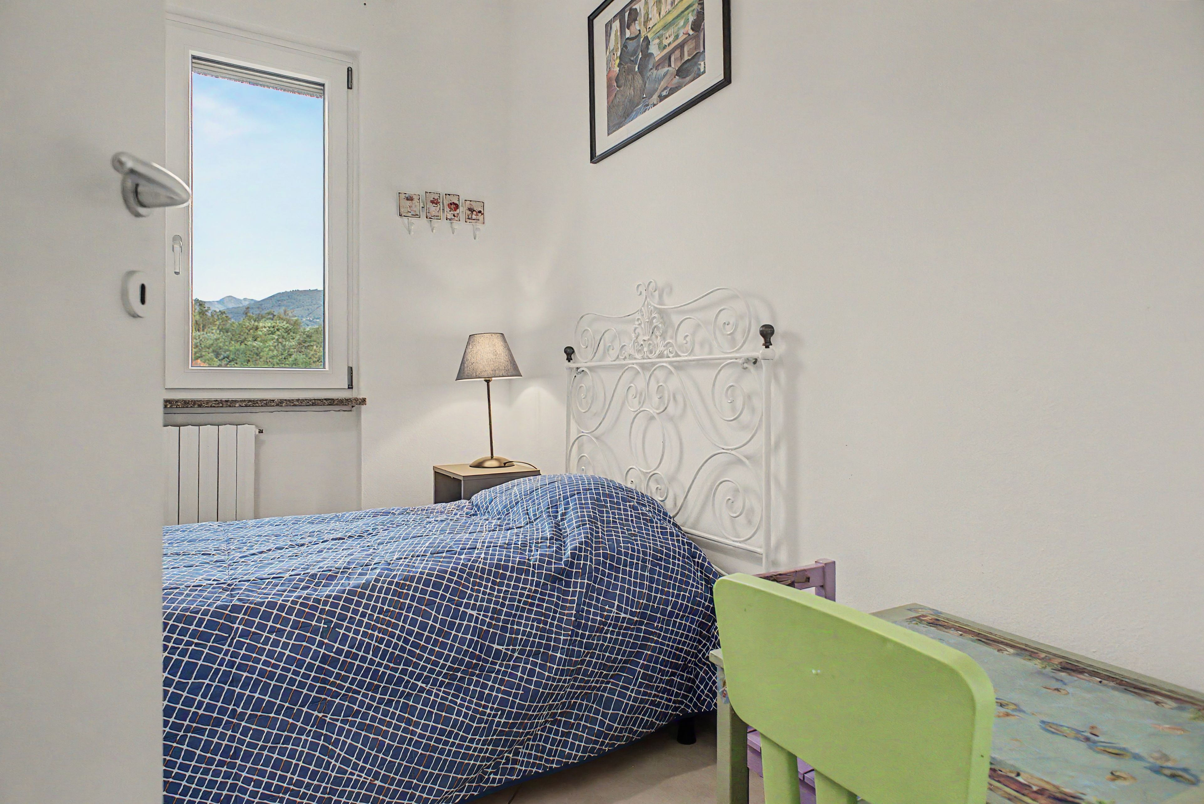 Ein Schlafzimmer mit einem Bett, einem Nachttisch, einer Lampe, einem Fenster mit Blick und einem gerahmten Bild an der Wand.