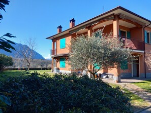 Ein zweistöckiges Gebäude mit Balkon, umgeben von Grünpflanzen und einem Berg im Hintergrund.