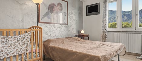A bedroom with a wooden bed, a bedside table with a lamp, a framed picture on the wall, an air conditioner, and a window with a view of the outdoors.