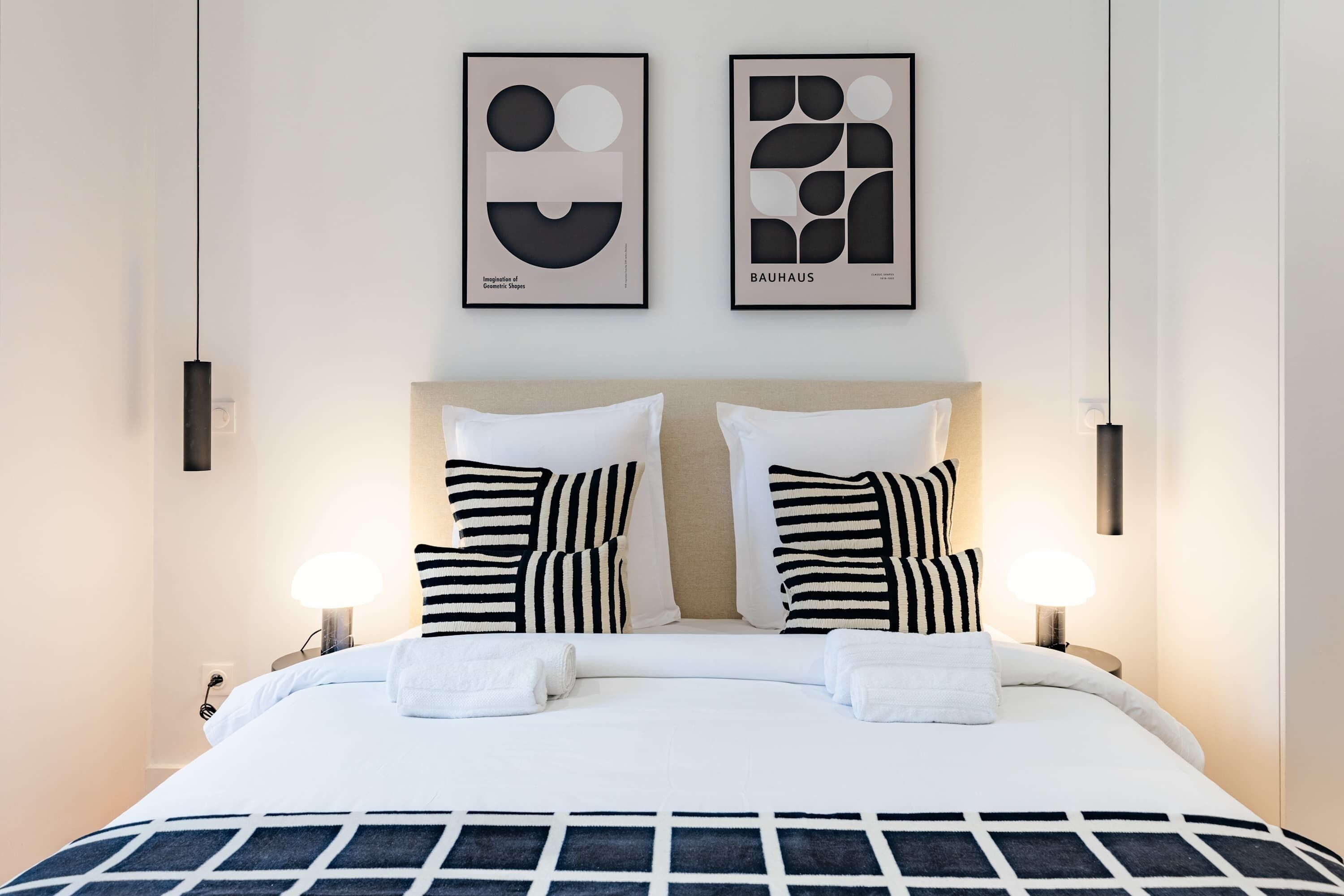 A modern hotel room with a large bed, two black and white striped pillows, a geometric patterned bedspread, and two framed abstract art pieces on the wall.