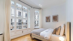 A hotel room with a bed, a bedside table with a lamp, and large windows with a view of the city. - Appartement de prestige climatisé (Paris)