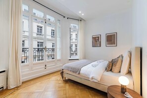 A hotel room with a bed, a bedside table with a lamp, and large windows with a view of the city. - Appartement de prestige climatisé (Paris)
