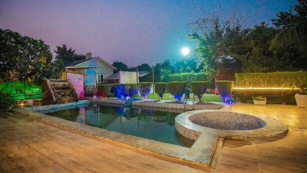 A swimming pool with a tiled edge, surrounded by a landscaped garden with various plants and a small house in the background. - Taj Royal Farm (Noida)