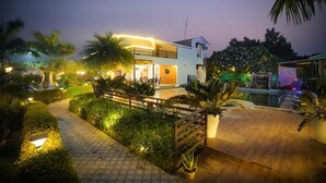 A modern two-story building with a well-lit entrance and a pool area. - Taj Royal Farm (Noida)