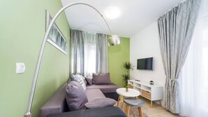Een moderne woonkamer met een hoekbank, een klein rond tafeltje, een aan de muur bevestigde televisie en een groot raam met gordijnen.