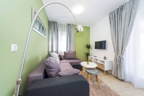 A modern living room with a sectional sofa, a small round table, a TV mounted on the wall, and a large window with curtains. - Apartment Maras 1 Zadar (Zadar)