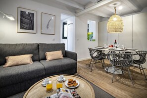 Ein modernes Wohnzimmer mit einem grauen Sofa, einem Esstisch mit Stühlen und einer Frühstücksecke mit Orangensaft und Gebäck.