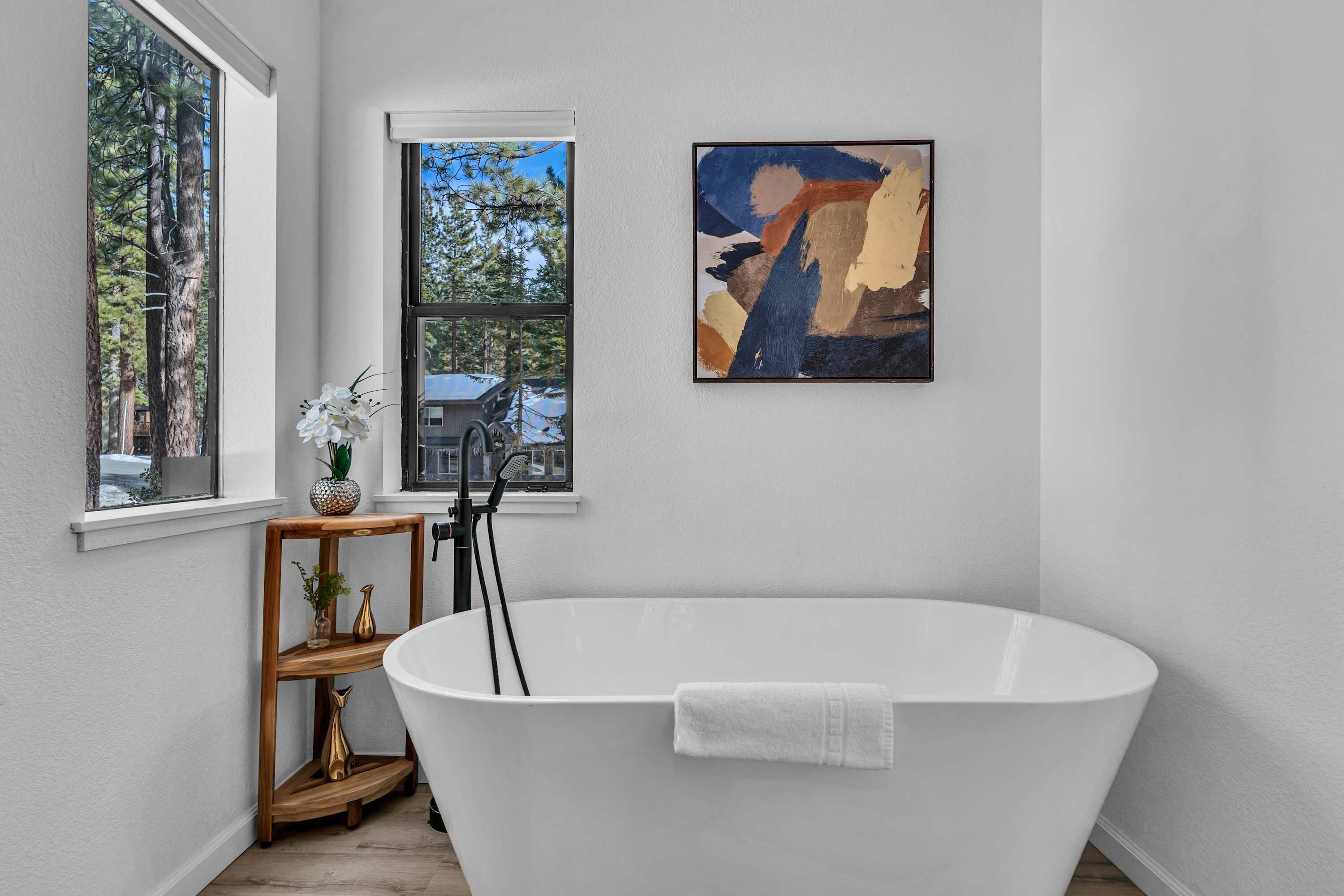 A modern bathroom with a freestanding bathtub, a wooden shelf with decorative items, and a painting on the wall.