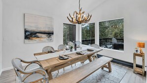 A dining room with a wooden table, chairs, a chandelier, and large windows with a view of trees. - Tahoe Retreat | Private Hot Tub & Scenic Views (Incline Village)