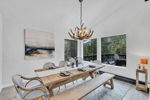 Comedor con mesa de madera, sillas, candelabro y ventanas amplias con vista a árboles.