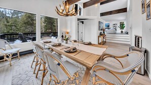 A spacious dining area with a wooden table set for six, surrounded by wicker chairs. A large chandelier hangs from the ceiling, and the room features exposed wooden beams and large windows offering a view of a deck and forest. - Tahoe Retreat | Private Hot Tub & Scenic Views (Incline Village)