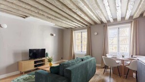 A modern living room with a green sofa, wooden flooring, a TV on a wooden cabinet, and a dining area with a table and chairs. - Charmant Appartement Climatisé - Marais (Paris)