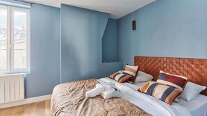 A bedroom with a blue wall, a wooden headboard, and a window with a view of buildings. - Charmant Appartement Climatisé - Marais (Paris)