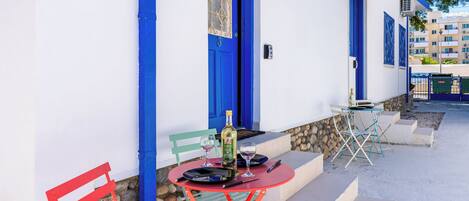 A white building with blue doors and windows, a red table with a bottle and glasses, and a green chair.