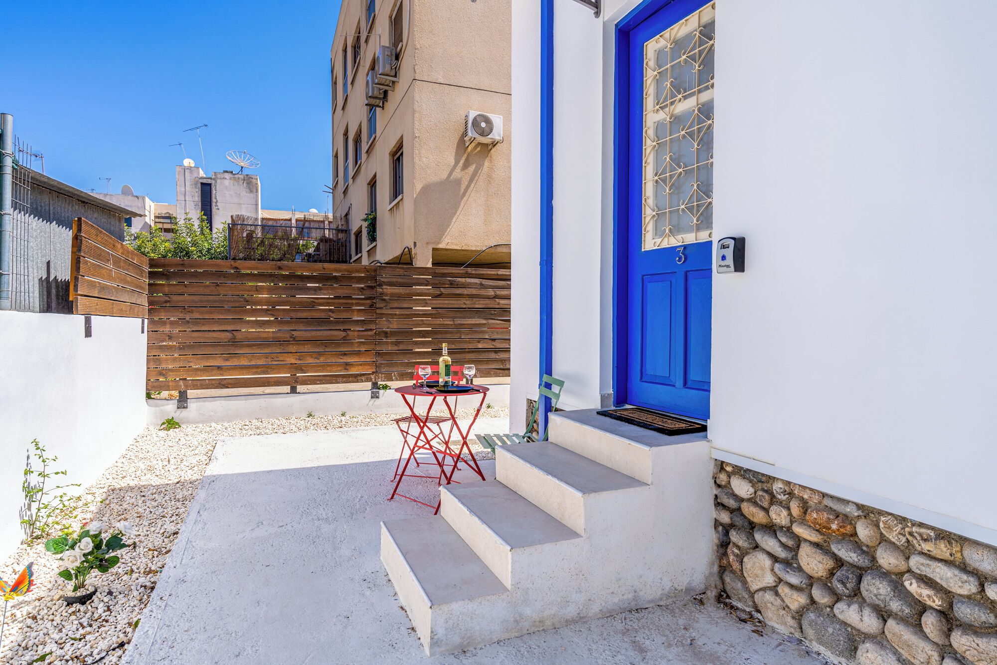 Una pequeña zona de estar al aire libre con una mesa y sillas, una puerta azul y un muro de piedra.