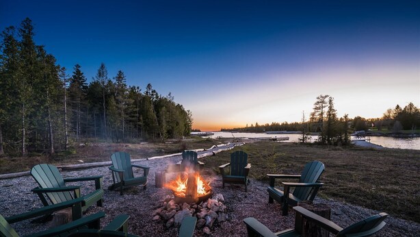 A fire pit surrounded by Adirondack chairs, a forested area, and a body of water.