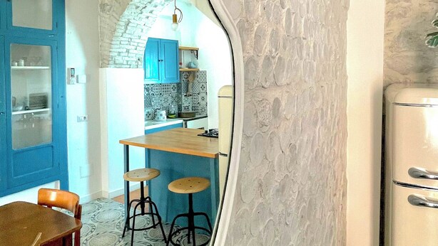 A room with a stone-textured wall, a mirror reflecting a dining area with a table and chairs, and a refrigerator.