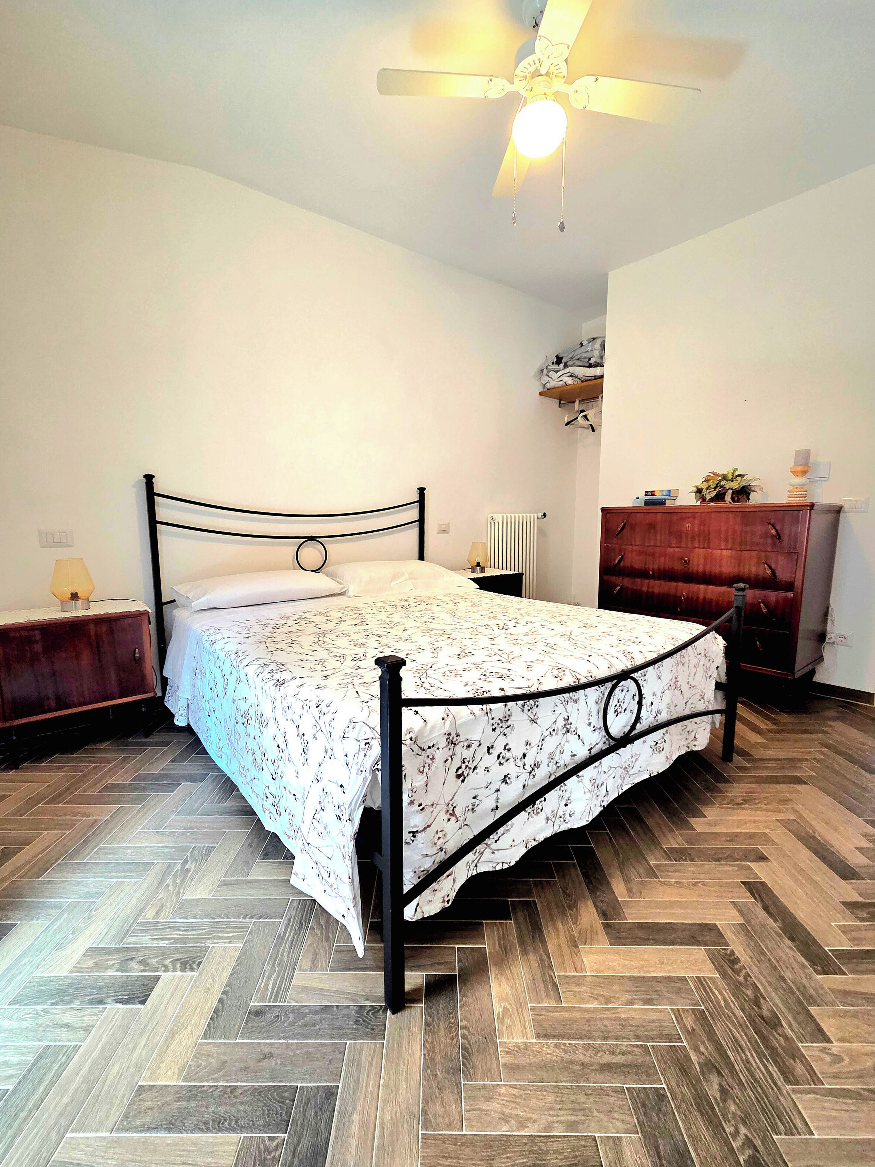 Een slaapkamer met een bed, een houten dressoir, een nachtkastje, een radiator en een plafondventilator.