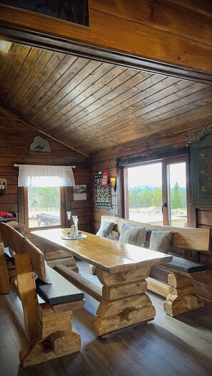 A rustic cabin interior with a wooden bench seating area around a central table, a window with a view of greenery, and a decorative wall hanging.