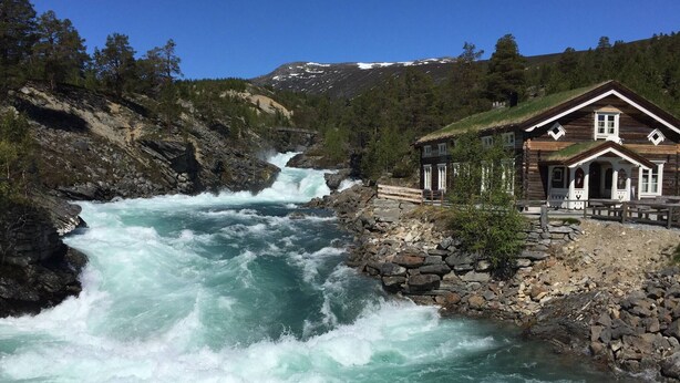 A wooden cabin with a thatched roof, located beside a turbulent river.