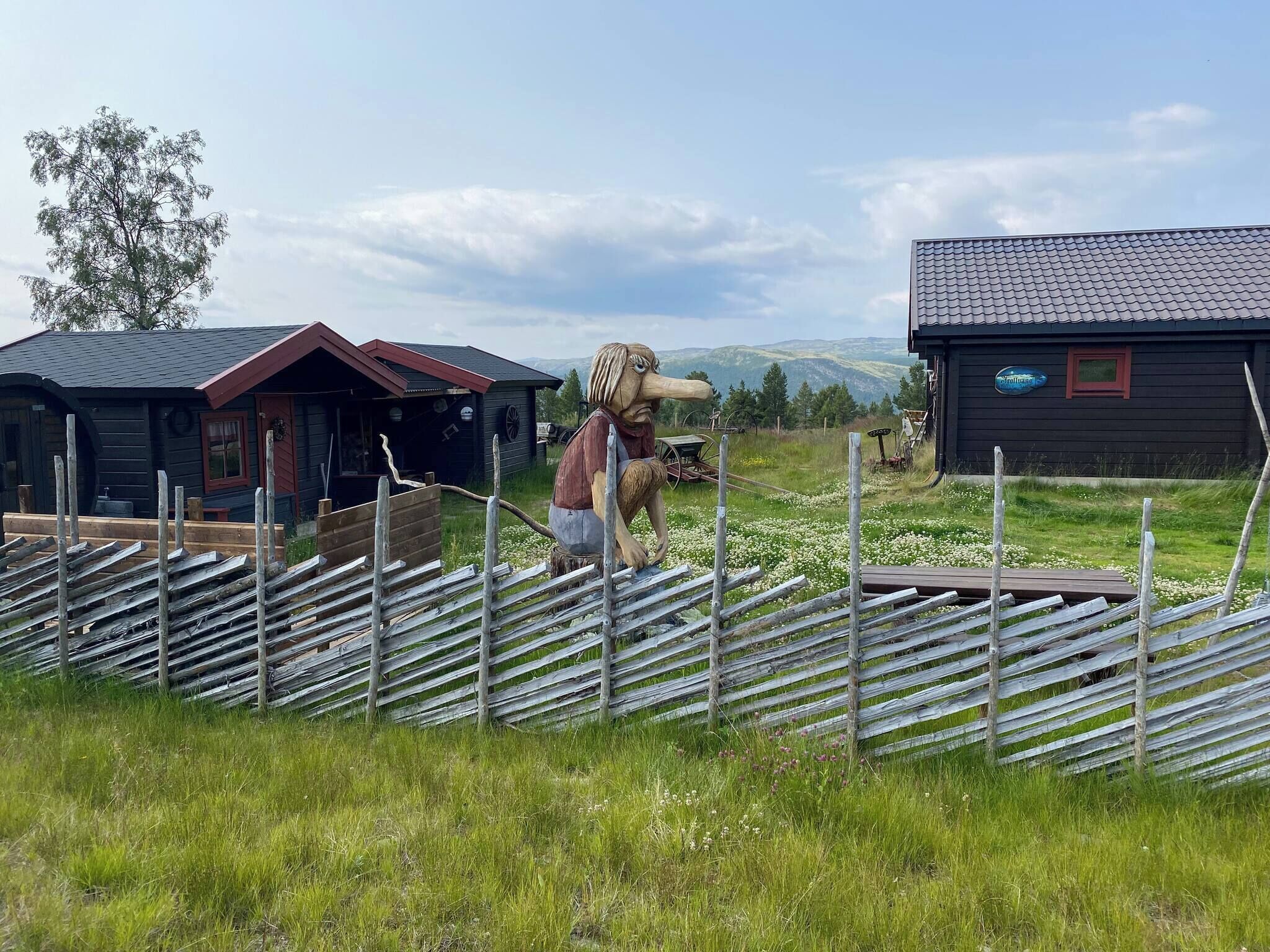A rustic wooden cabin with a sloped roof, surrounded by a wooden fence and a field of white flowers.