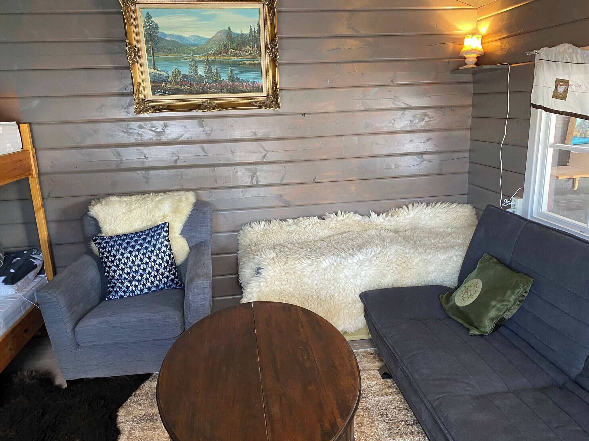 Ein gemütliches Wohnzimmer mit einer Holzwand, einem gerahmten Landschaftsbild, einem grauen Sofa, einem blauen Sessel, einem runden Holztisch und einem flauschigen weißen Teppich.