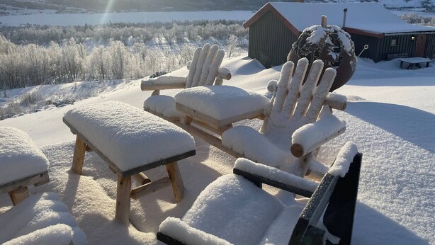 Un'area salotto esterna coperta di neve con poltrone Adirondack e un tavolo, con vista su un paesaggio montuoso e un cottage in legno sullo sfondo.