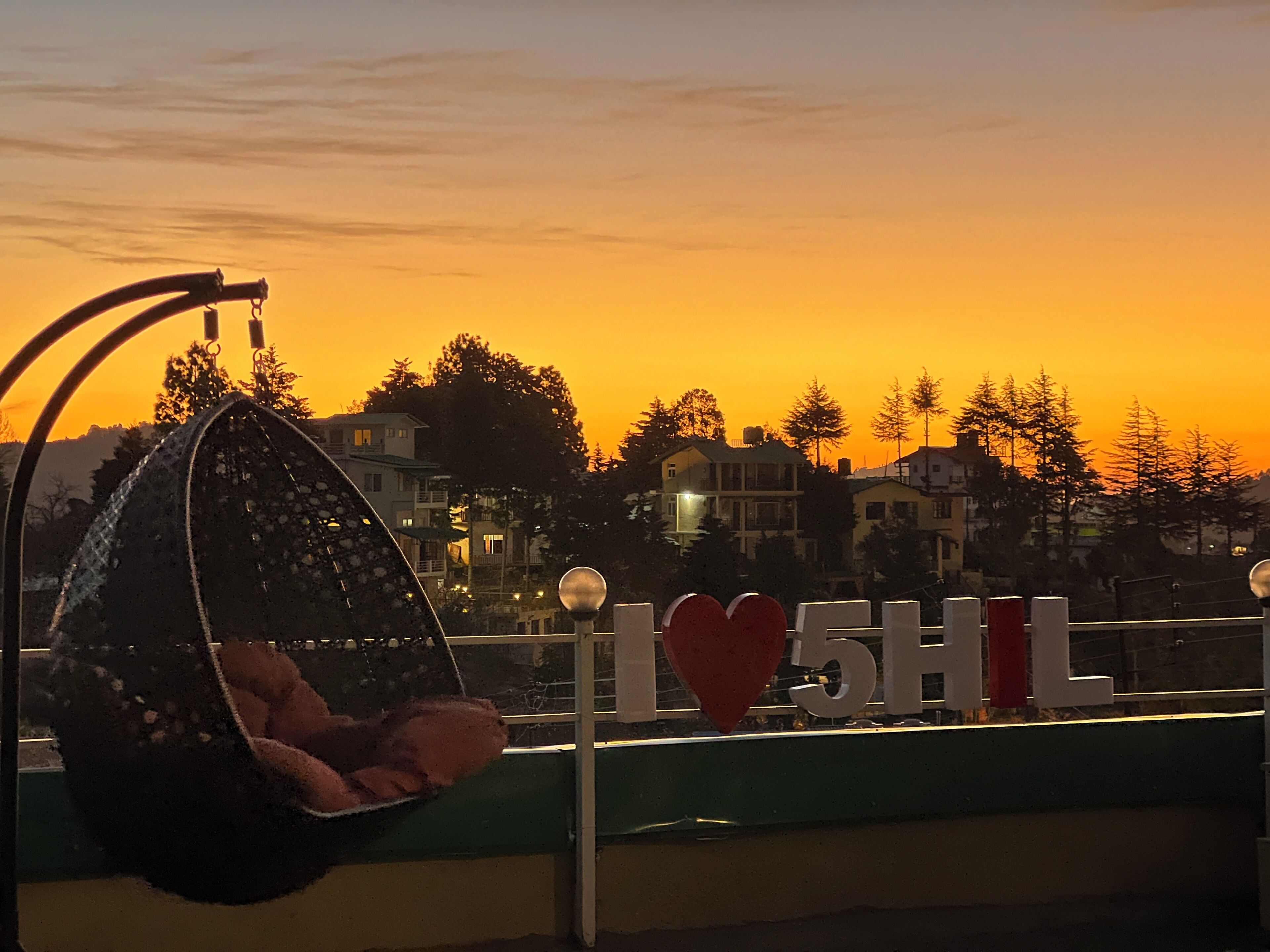 Une chaise suspendue avec un coussin, une balustrade et une pancarte portant l’inscription « I ♥ 5HL », sur un balcon donnant sur un quartier résidentiel, au coucher du soleil.