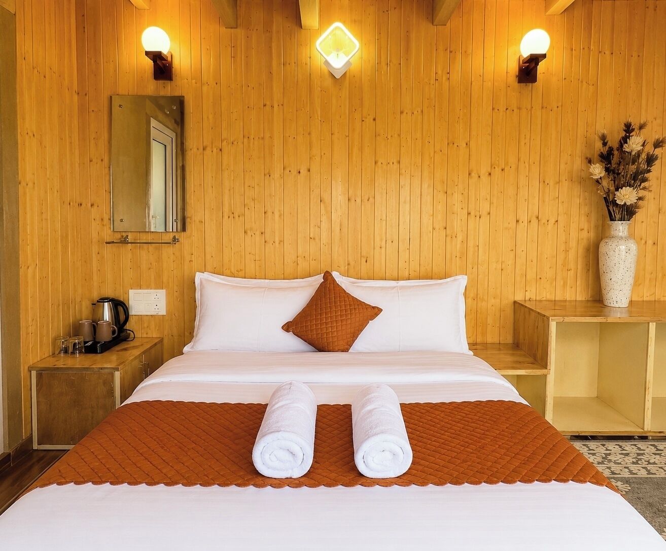 A hotel room with a wooden paneled wall, a bed with white linens, a brown quilted pillow, and two rolled towels.