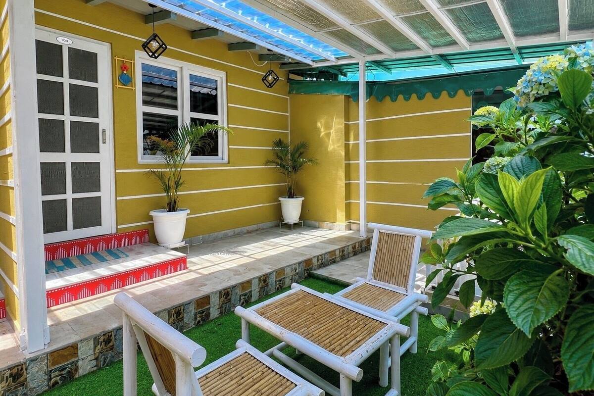 A yellow building with a covered patio, two white chairs, and potted plants.