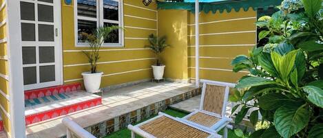 A yellow building with a covered patio, two white chairs, and potted plants.
