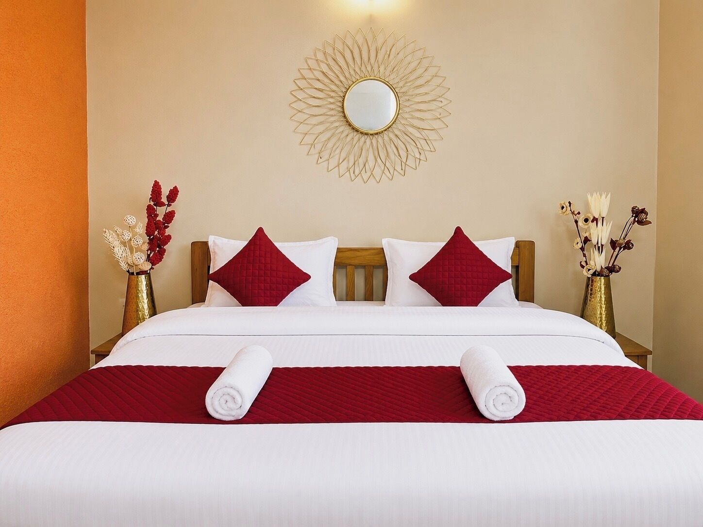 A neatly made bed with white and red bedding, a round mirror on the wall, and two vases with flowers on either side.