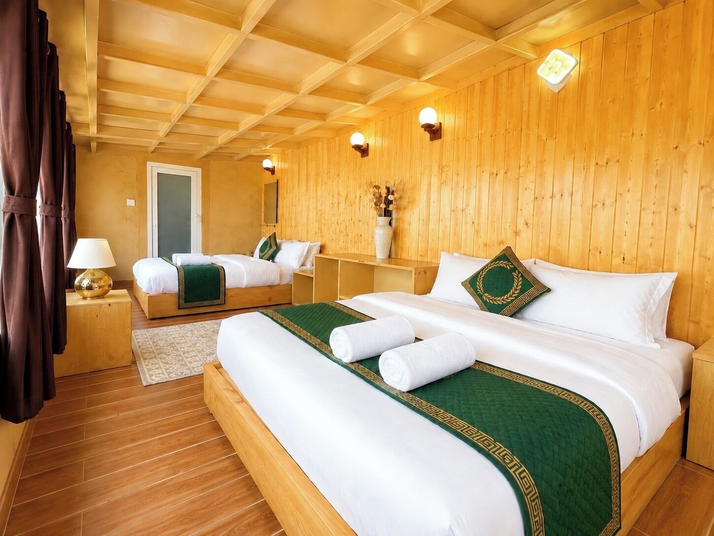 Habitación de hotel con paredes y techo de madera, cama con ropa de cama blanca y detalles en verde, mesita de noche con lámpara y una pequeña mesa con un florero.