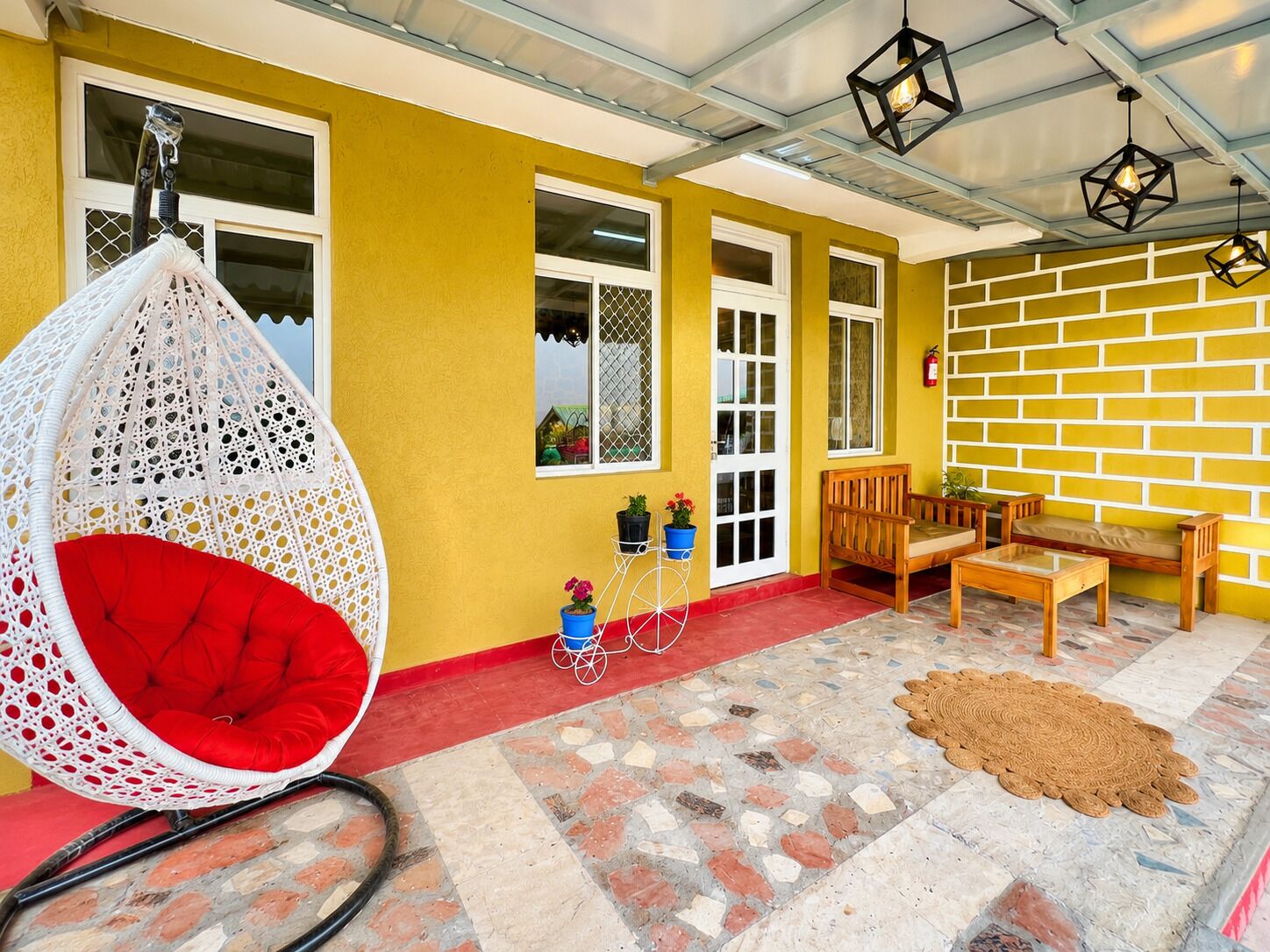 A yellow exterior wall, a white hanging chair with red cushions, a wooden bench, and a small table with potted plants.